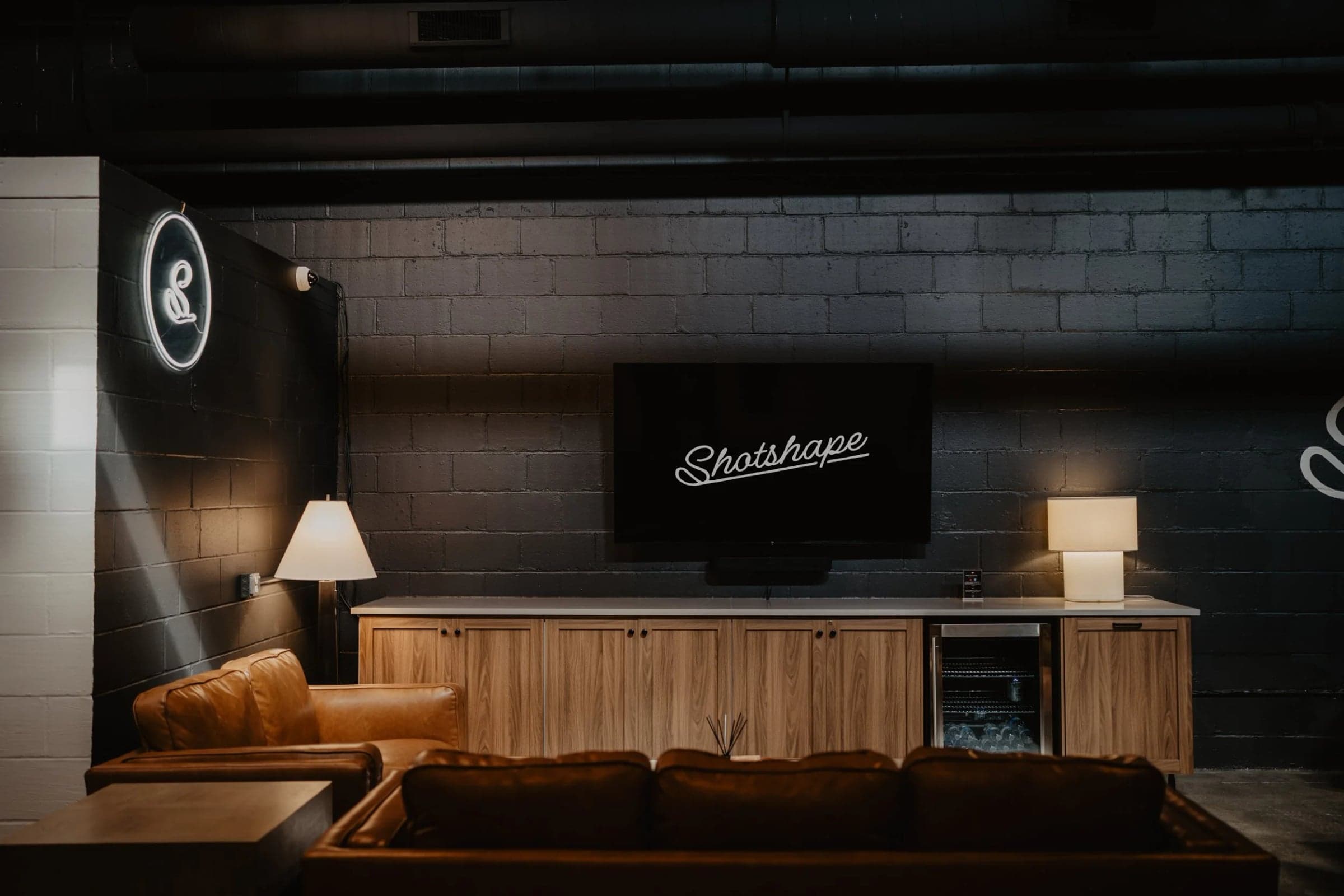 Shotshape Golf Studio - Northeast — Lounge end of the studio: painted black brick, deep tan leather sofa, timber credenza with