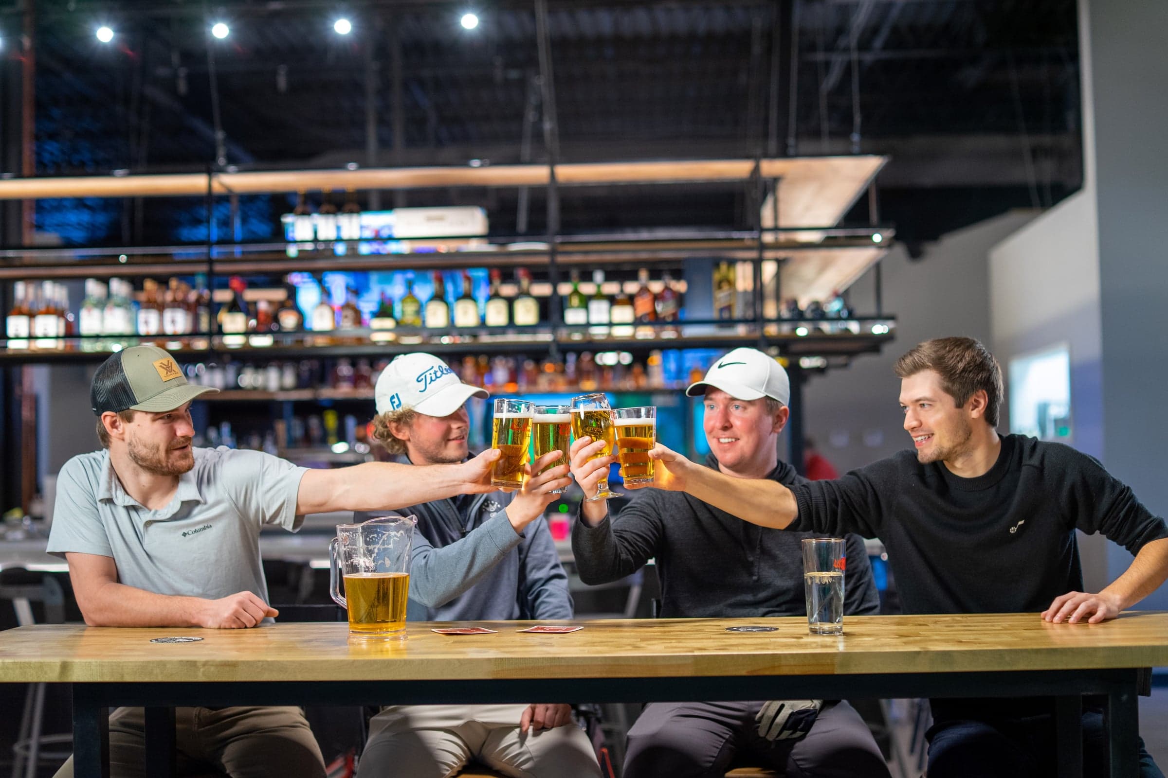 Element Indoor Golf - Burnsville — Four patrons toasting beers at a butcher-block high table, backlit bar and bay screens fil